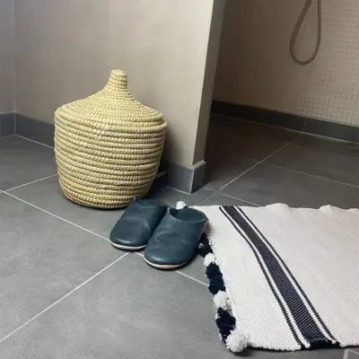 Marokkanischer handgewebter Badeteppich in Weiß mit zwei schwarzen Streifen mit weißen und schwarzen Pompons, auf dem Badezimmerboden vor der Duschkabine liegend, nah