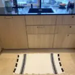 Moroccan handwoven bath mat in white with two black stripes with white and black pompoms, lying on the kitchen floor in front of the sink