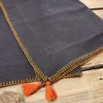 To koksgrå marokkanske håndbroderede dækkeservietter med orange kant og orange pomponer, foldet oven på hinanden, tæt