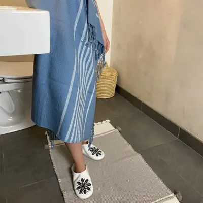 Model wearing Moroccan handwoven hammam towel in blue with white slippers on, outside in a bathroom