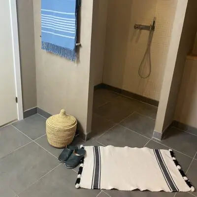 Marokkanische handgewebte Badematte in Weiß mit zwei schwarzen Streifen mit weißen und schwarzen Pompons, auf dem Badezimmerboden vor der Duschkabine liegend
