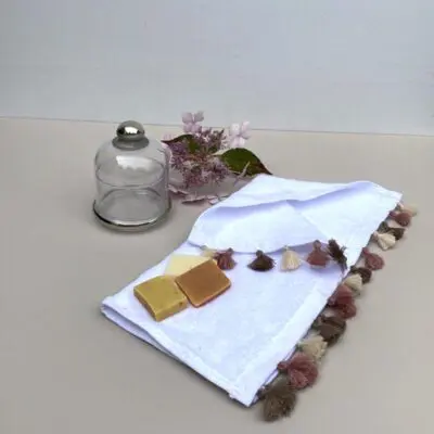 Kleines weißes handgewebtes Handtuch mit ockerfarbenen Pompons, darauf Seifen und daneben ein Glasgefäß