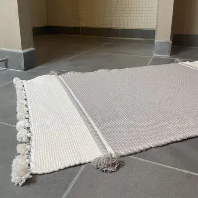 Marokkansk håndvævet bademåtte i grå med hvide og grå pomponer, liggende på badeværelses gulv foran brusekabinen, tæt