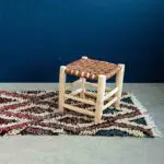 Tabouret marocain en bois fait main avec siège en cuir tressé, posé sur un tapis avec un mur bleu derrière