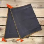 Two charcoal gray Moroccan hand-embroidered placemats with orange border and orange pom-poms, folded on top of each other