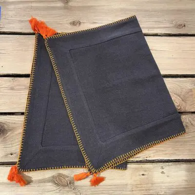 Two charcoal gray Moroccan hand-embroidered placemats with orange border and orange pom-poms, folded on top of each other