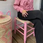 Model sitting on Moroccan handmade stool in pink, wearing pink shirt with pink basket next to it