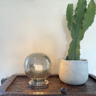 Marokkaanse handgemaakte tafellamp van zilver metaal met eenvoudig gatenpatroon, verlicht op een plank naast de plant
