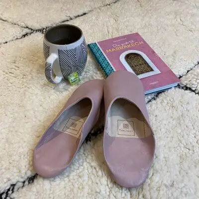 Moroccan handmade slippers in light violet on top of Beni Ouarain rug with Marrakech guide and mug next to it