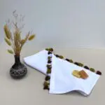 Small white handwoven towel with ocher colored pompoms, with soaps on top and a plant next to it
