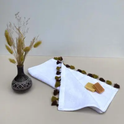 Petite serviette blanche tissée à la main avec des pompons de couleur ocre, avec des savons dessus et une plante à côté
