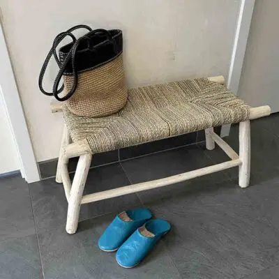 Moroccan handmade wooden bench with wicker raffia seat, standing in entrance hall, with a bag on top and slippers in front