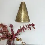 Håndlavet væglampe i guldmetal med marokkansk mønster hængende på hvid væg med blomster under