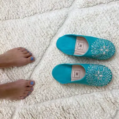 Marokkaanse handgemaakte pantoffels in turquoise met wit patroon bovenop beni ouarain tapijt met voetmodel ernaast