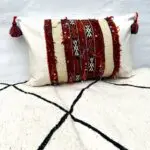 Housse de coussin boho kilim vintage tissée à la main en rouge et beige avec motif marocain avec pompons sur les bords, posée sur un pouf