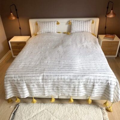 Hvidt marokkansk håndvævet sengetæppe med sorte striber og gule pomponer, på en seng med matchende puder