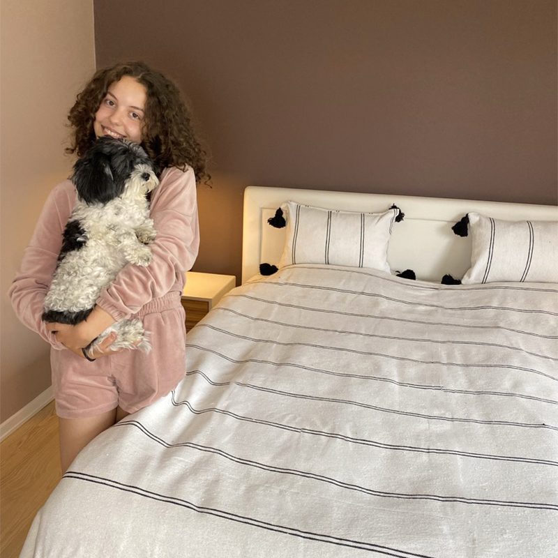 Hvidt marokkansk håndvævet sengetæppe med sorte striber og sorte pomponer på seng, med matchende puder og en model der holder en hund