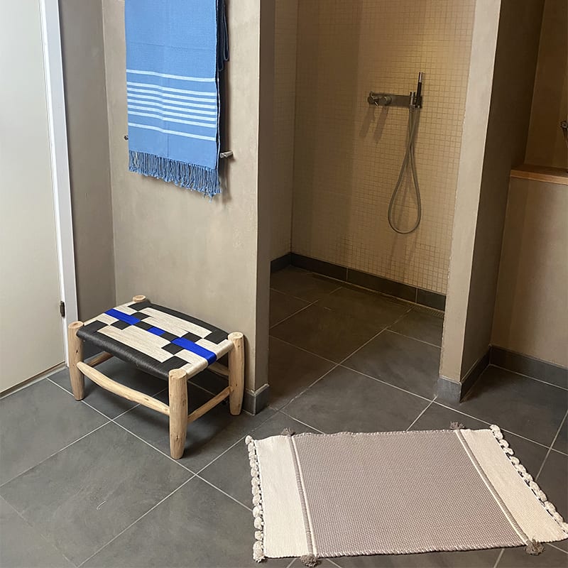 Marokkansk håndvævet bademåtte i grå med hvide og grå pomponer, liggende på badeværelses gulv foran brusekabinen