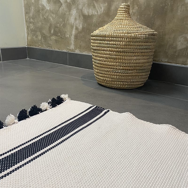 Marokkansk håndvævet bademåtte i hvid med to sorte striber med hvide og sorte pomponer, liggende på badeværelses gulv foran en håndflettet kurv