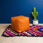 Håndvævet boucherouite tæppe i flerfarvet pile mønster, med læderpuf ovenpå, og plante ved siden af