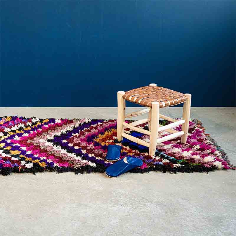 Håndvævet boucherouite tæppe i flerfarvet pile mønster, med træskammel og blå hjemmesko ovenpå