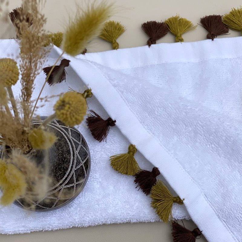 Lille hvidt håndvævet håndklæde med okker farvet pomponer, med en plante oven på