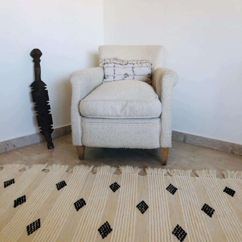 Håndvævet bomuldstæppe i hvid med marokkansk stribemønster og prikmønster i sort med hvide kvaster, foran hvid lænestol