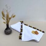 Lille hvidt håndvævet håndklæde med okker farvet pomponer, med sæber oven på og en plante ved siden af