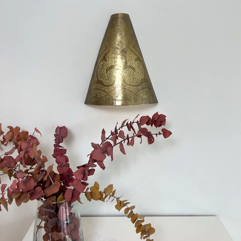 Håndlavet væglampe i guldtmetal med marokkansk mønster, hængende over en reol med en vase på