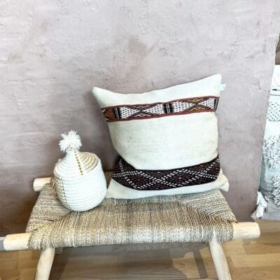 Handwoven vintage kilim montagne cushion cover in beige with Moroccan pattern in black, red and white standing on bench with basket next to it