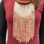 Pärlhalsband med halsring i ljusbrunt flätat läder och vita pärlor som dominerande färg. Orangea och gyllene pärlor blandas in och ger ett varmt och glödande uttryck, som påminner om saffran i snö.